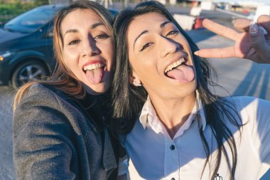 İki güzel Latin bayan arkadaş dillerini uzatıp cep telefonlarıyla selfie çekiyorlar. Arkadaşlık ve dostluk kavramı.