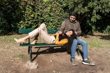 Kız arkadaşı dizlerinin üzerinde yatarken gülümseyen adam bankta oturuyor. Aşk konsepti.