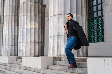 Siyah ceketli ve kot pantolonlu genç adam kolonları olan geleneksel bir taş binanın merdivenlerinden iniyor.