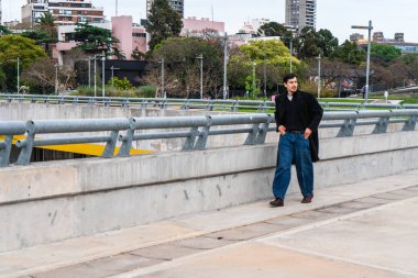 Buenos Aires 'te beton üstgeçit boyunca yürüyen genç adam, şehir manzarası ve yeşil park arka planının ortasında sıradan bir ceket ve kot pantolon giyiyor.
