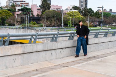 Buenos Aires 'te beton üstgeçit boyunca yürüyen genç adam, şehir manzarası ve yeşil park arka planının ortasında sıradan bir ceket ve kot pantolon giyiyor.