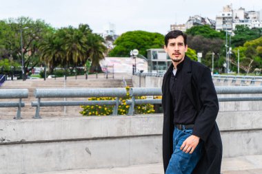 Siyah ceketli ve kot pantolonlu genç bir adam parka ve şehir binalarına bakan bir köprüde yürüyor.