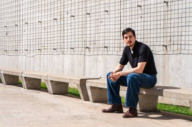 Sert beton bir bankta oturan genç adam, doğrudan kameraya bakıyor, günlük giysiler giyiyor.