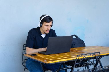 Kulaklık takan, dizüstü bilgisayarda yazan açık bir kafe masasında çalışan ya da ders çalışan genç bir adam.