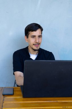 Uzaktan kumandayla uğraşan genç bir adam, tahta masada dizüstü bilgisayar kullanıyor. Dijital görevlere odaklanmış, esnek çalışma alanının keyfini çıkarıyor