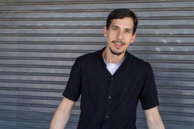 Genç adam gülümsüyor, kendine güven ve mutluluk gösteriyor. Şehir ortamında metal bir kapının önünde dikiliyorum.