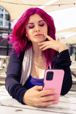 Pembe saçlı ve makyajlı genç bir kadın açık havada pembe bir akıllı telefon kullanırken beyaz bir masada oturuyor.