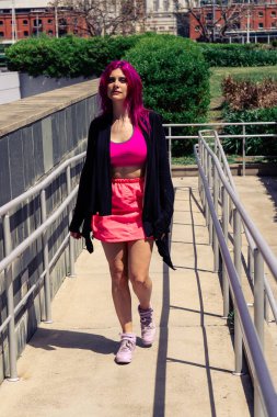 Neşeli pembe saçları ve dövmeleri olan kendine güvenen genç bir kadın güneşli bir yaz gününde şehir rampasında yürüyor, şık ve cesur.