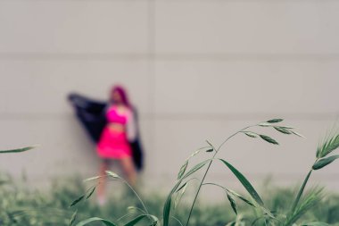 Çimenler, açık pembe giymiş bulanık bir kadınla soluk gri bir duvara karşı çelişiyor ve şehir gizemi yaratıyor.