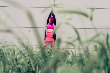 İsyankar, pembe saçlı ve dövmeli bir kadın modern bir şehir duvarının önünde uzun otların arkasında poz veriyor.