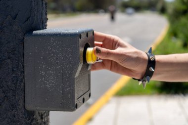 Elinde çivili bir bilezik, sarı bir yaya geçidi düğmesine basıyor ve caddeyi geçmeyi bekliyor.