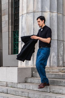 Büyük bir binanın yanındaki taş merdivenlerden inen bir adam şehir ortamında siyah bir ceket taşıyor.