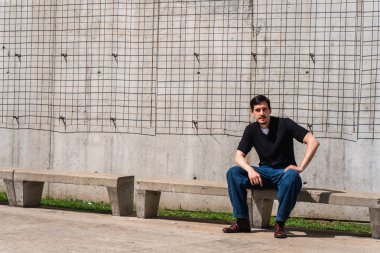 Genç adam beton minimalist beton bir duvara karşı düz bir bankta oturuyor. Açık hava ortamında metal bir ızgara var.