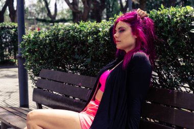 Parlak, pembe saçlı genç bir kadın güneşli bir günde ahşap bir bankta oturuyor, dinleniyor ve kafa yoruyor.