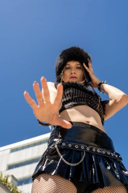 Alternatif punk-goth kıyafetli bir kadın açık mavi gökyüzüne el kaldırıyor, kendine güvenen asi cesur bir açıklama yapıyor.