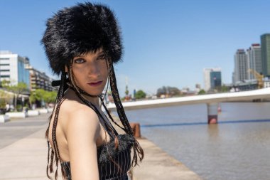 Genç adam kameraya bakıyor, siyah kürklü ushanka şapkalı ve örgülü, Buenos Aires 'te nehrin kenarında duruyor.