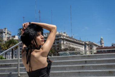 Şehir merdivenlerinde poz veren, kürk şapkalı ve çizgili üstlü güneşli bir günde arka planda şehir binaları olan genç bir kadın.