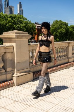 Siyah makyajlı, tüylü çoraplı ve mini etekli alternatif modaya sahip genç bir adam şehir ortamında yürüyor.