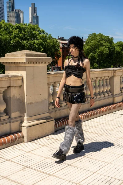 Siyah makyajlı, tüylü çoraplı ve mini etekli alternatif modaya sahip genç bir adam şehir ortamında yürüyor.