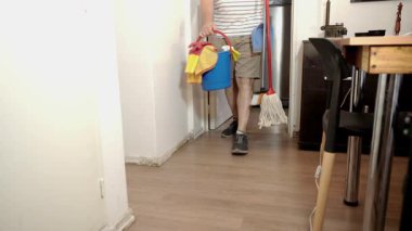 Beyaz bir adam bir paspas ve bir kova temizlik malzemesi taşıyor. Onları bir köşeye koyar, ev işlerini bitirir.