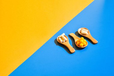 Ahşap kaşıkta haplar mavi ve sarı arka planda. Sert ışık ve gölgeler. Modern izometrik yaratıcı minimalizm kavramı. Vitaminler, prebiyotikler, probiyotikler. Sonbahar vitamin dozu.