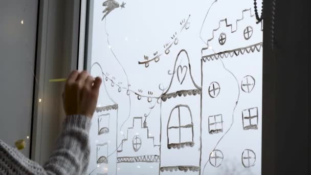 Vrouwelijke hand tekent kerstvakantie decoratie op glas. Nieuwjaarsdecor. stadsgebouwen bloemenslingers lichten. hygge, decoratie en kerstconcept — Stockvideo