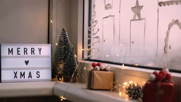 Lightbox con testo Buon Natale perde sul davanzale della finestra. Decorazioni di anno nuovo, luci di ghirlanda e alberi di Natale. Finestra dipinta con spray neve. — Video Stock