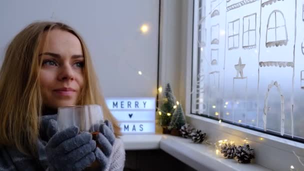 Jovencita sentada junto a la ventana y bebiendo té. Concepto casero de Hygge. Feliz Navidad, luz de guirnalda, quédate en casa. Feliz año nuevo 2021. Paisaje de vacaciones de invierno. — Vídeo de stock