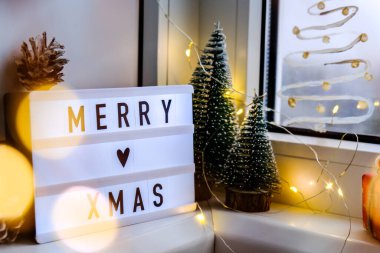 Pencere pervazında Merry XMAS yazan ışık kutusu. Yeni yıl süslemeleri, çelenk ışıkları ve Noel ağaçları. Pencere kar spreyi ile boyanmış. Hygge evde.