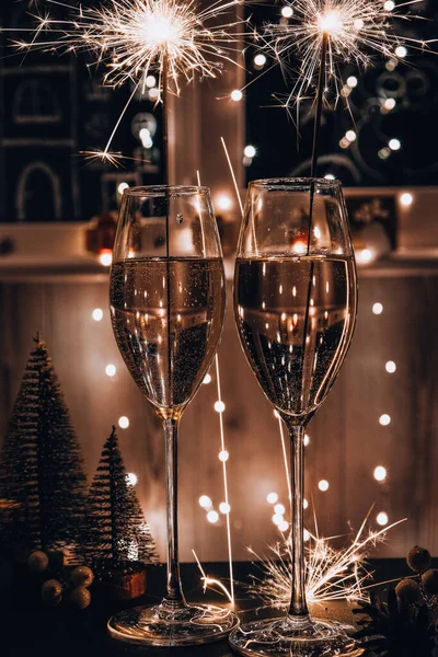 Two glasses of sparkling wine champagne with sparklers. Dark background with yellow light bokeh. Christmas tree toys on the table. New Year\'s bengal fire. Christmas winter holidays
