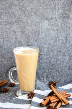 Sütlü, tarçınlı ve anason yıldızlı cam bardakta geleneksel kış içkisi gri bir bezin üzerinde krem şanti, kumaş arka planında tarçın çubukları. Kafe menüsü reklamı. Kahve.