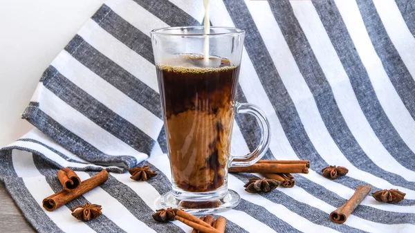 Pouring of milk into glass with cold coffee on grey table with cinnamon sticks and anise stars, Coffee with ice and milk, brown table, selective focus