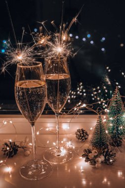 İki bardak köpüklü şampanya ve maytap. Sarı ışık saçan koyu bir arka plan. Masada Noel ağacı oyuncakları var. Yeni yılın Bengal ateşi. Noel kış tatili