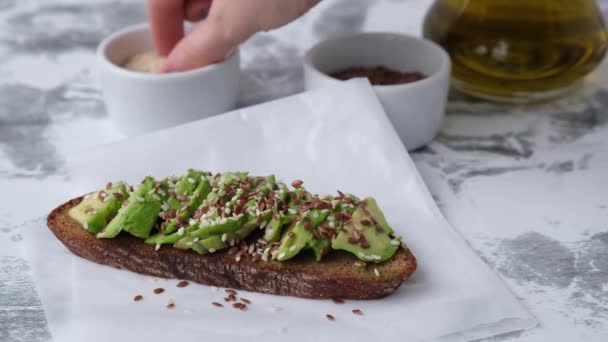 Ženská ruka dělá zdravé avokádové toasty na dřevěné desce. Sezamová a lněná semena. Vegetariánské jídlo. — Stock video
