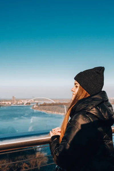 Young beautiful woman travelling. Staying on the bridge against blue sky and river in Kiev Ukraine. Cold weather clothes. Happy blogger traveller.