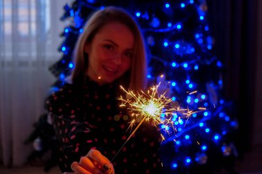 Noel ağacının yanında bir bardak şampanya ve bengal ışığıyla oturan mutlu genç kadın. Yeni yıl partisi kutlaması. Noel kış tatili. Yeni Yıl Arifesi.