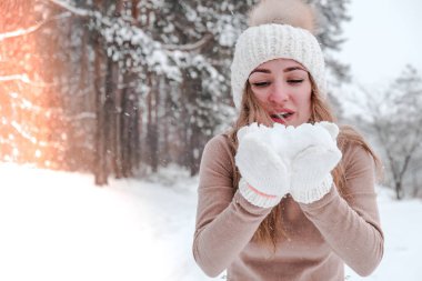 Noel, tatil ve sezon konsepti. Genç mutlu kadın kış ormanlarında kar üflüyor. Sıcak giysi, eldiven ve şapka..