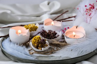 Paskalya bayramı. Kendiniz yapın. Moda Paskalya dekorasyonları. Tabağında kuru çim çiçekleri olan organik yumurtalar. Yumurtaların beyaz kabuğunda mum ışığı. Kuş tüyü. Uzayı kopyala Paskalya kartı. Pastel renkler.