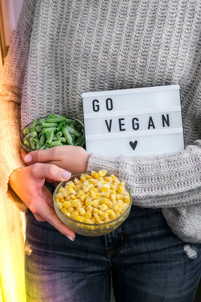 Lightbox with text GO VEGAN in female hands. Veganism, vegetarian healthy lifestyle. Frozen food in bowl. Healthy eating vegan. Green beans and Yellow corn