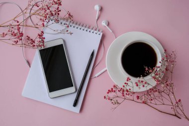 Kulaklıklı cep telefonlu boş bir defter aç. Sade kahve ve ekran. Kuru pembe çiçek süslemesi. İş yeri. Uzayı metin için kopyala. Üst Manzara. Pastel renkler