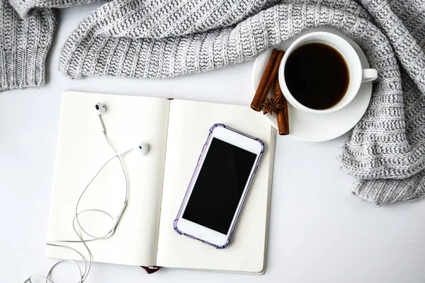 Cup of coffee with notebook phone headphones cinnamon sticks and anise star on white background. Sweater around. Winter morning routine. Coffee break. Copy space. Top view. Breakfast. Flat lay