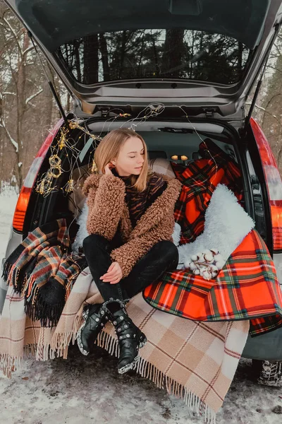 Blonde woman wrapped in blanket in trunk car. Travel in winter. Car decorated with festive Christmas lights. Outdoor picnic