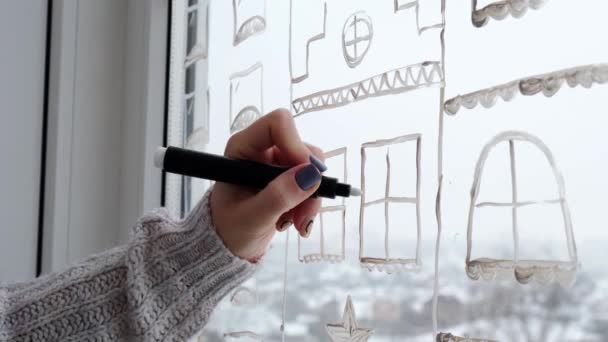 La mano femenina dibuja la decoración de las fiestas de Navidad en el cristal de la ventana. Paisaje de Año Nuevo. edificios de la ciudad guirnaldas luces. hygge, decoración y concepto de Navidad — Vídeos de Stock