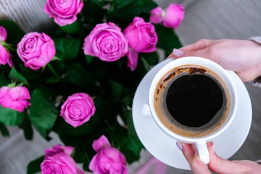 Bir fincan kahve ve pembe güller. Tatil konsepti, doğum günü için bir buket gül, sabah kahvaltısında bir buket çiçek. Kapuçino tutan kadın elleri var. Sevgililer Günü