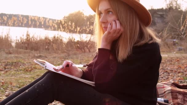 Mujer rubia caucásica de cámara lenta con sombrero beige en suéter negro escribir en el cuaderno en el campo. Hora dorada, casa de campo. Viajes locales. Una vida lenta. Salud mental — Vídeos de Stock