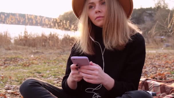 スローモーション黒セーターでベージュの帽子を持つ白人ブロンドの女性は電話を使用し、田舎で音楽のヘッドフォンを聞きます。黄金の時間、コッタゴコア。地方旅行。遅い生活。精神衛生 — ストック動画