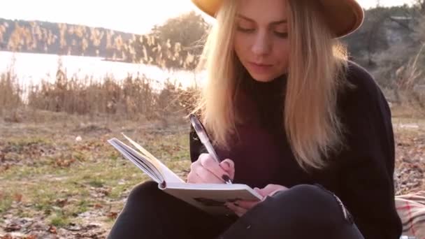 4k Mujer rubia caucásica con sombrero beige en suéter negro escribir en el cuaderno en el campo. Hora dorada, casa de campo. Viajes locales. Una vida lenta. Salud mental — Vídeos de Stock