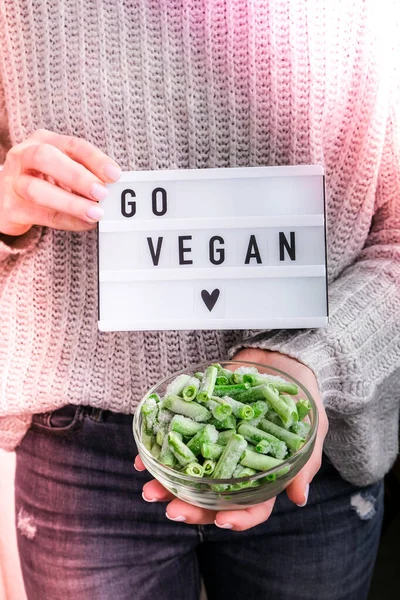 Lightbox with text GO VEGAN in female hands. Veganism, vegetarian healthy lifestyle. Frozen food in bowl. Healthy eating vegan. Green beans