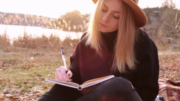 Femme blonde caucasienne au mouvement lent avec chapeau beige en pull noir écrire sur carnet à la campagne. Heure d'or, Cotagecore. Voyage local. Une vie lente. Santé mentale — Video