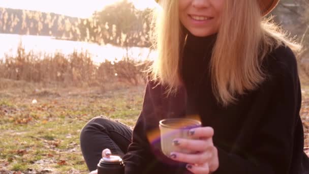 Mujer rubia caucásica de cámara lenta con sombrero beige en suéter negro bebe té caliente del termo en el campo. Hora dorada, casa de campo. Viajes locales. Una vida lenta. Salud mental — Vídeos de Stock